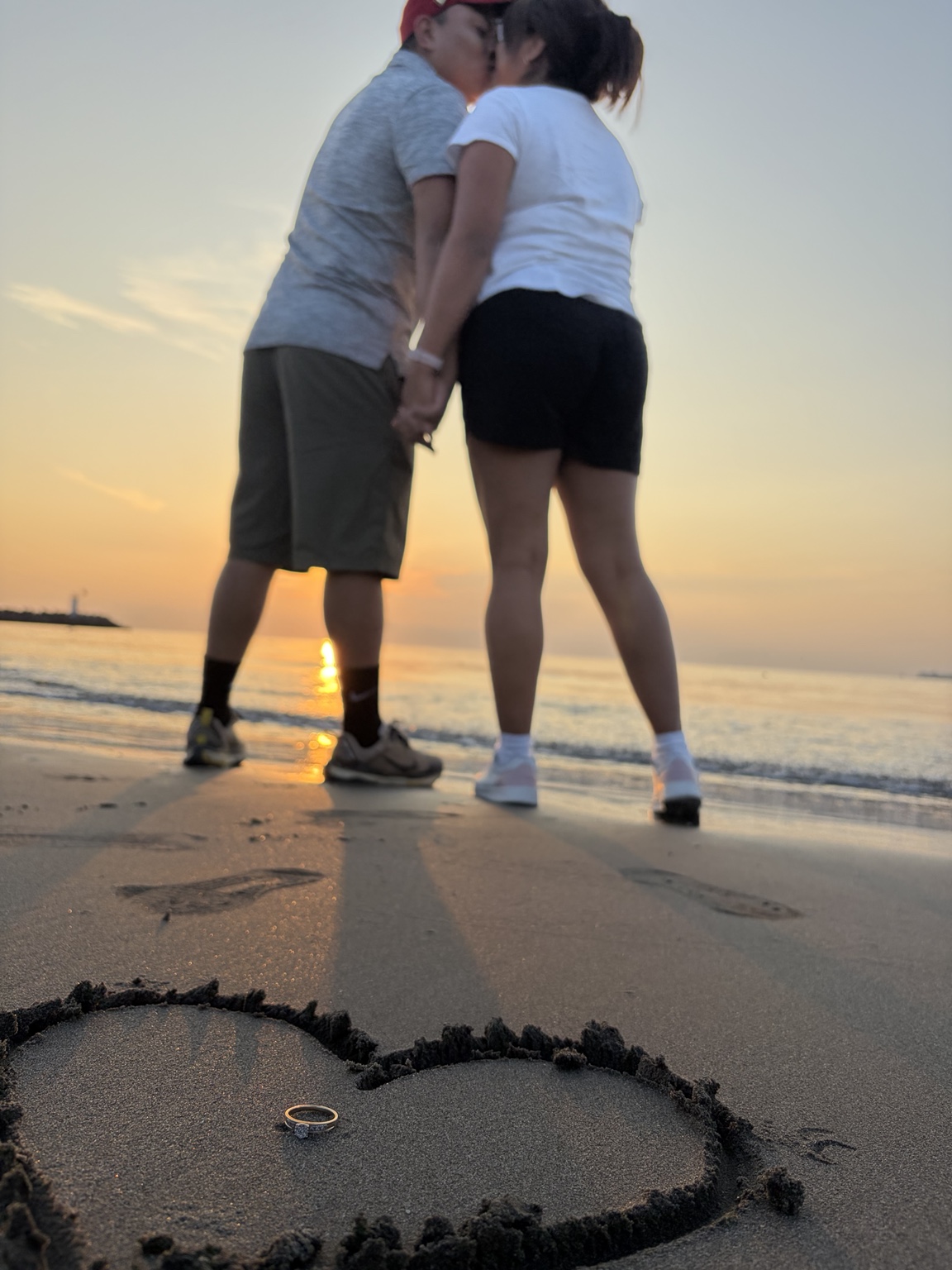 Felipe y Belen en la playa al atardecer, con un corazón y un anillo en la arena